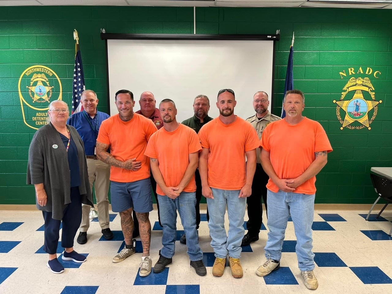 group photo posed in front of an American flag and the logo of Northwestern Adult Correctional Center; in the foreground, four smiling men wearing jeans and bright orange T-shirts. In the back row, four correctional officers in uniform or business attire. At the side, a smiling grey-haired woman, the adult education instructor