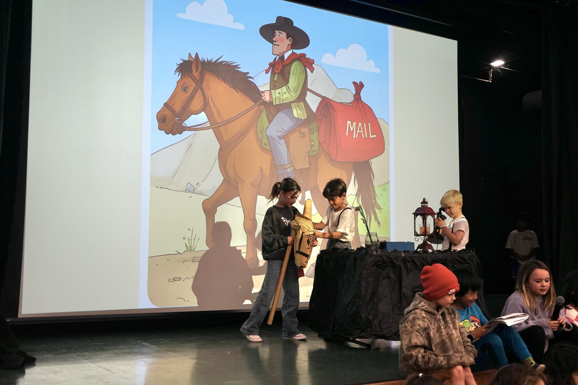 Two students stand at a table using an old phone. One student is riding a stick horse. An image of a man on a horse in western clothes and a mail bag is projected behind them. 