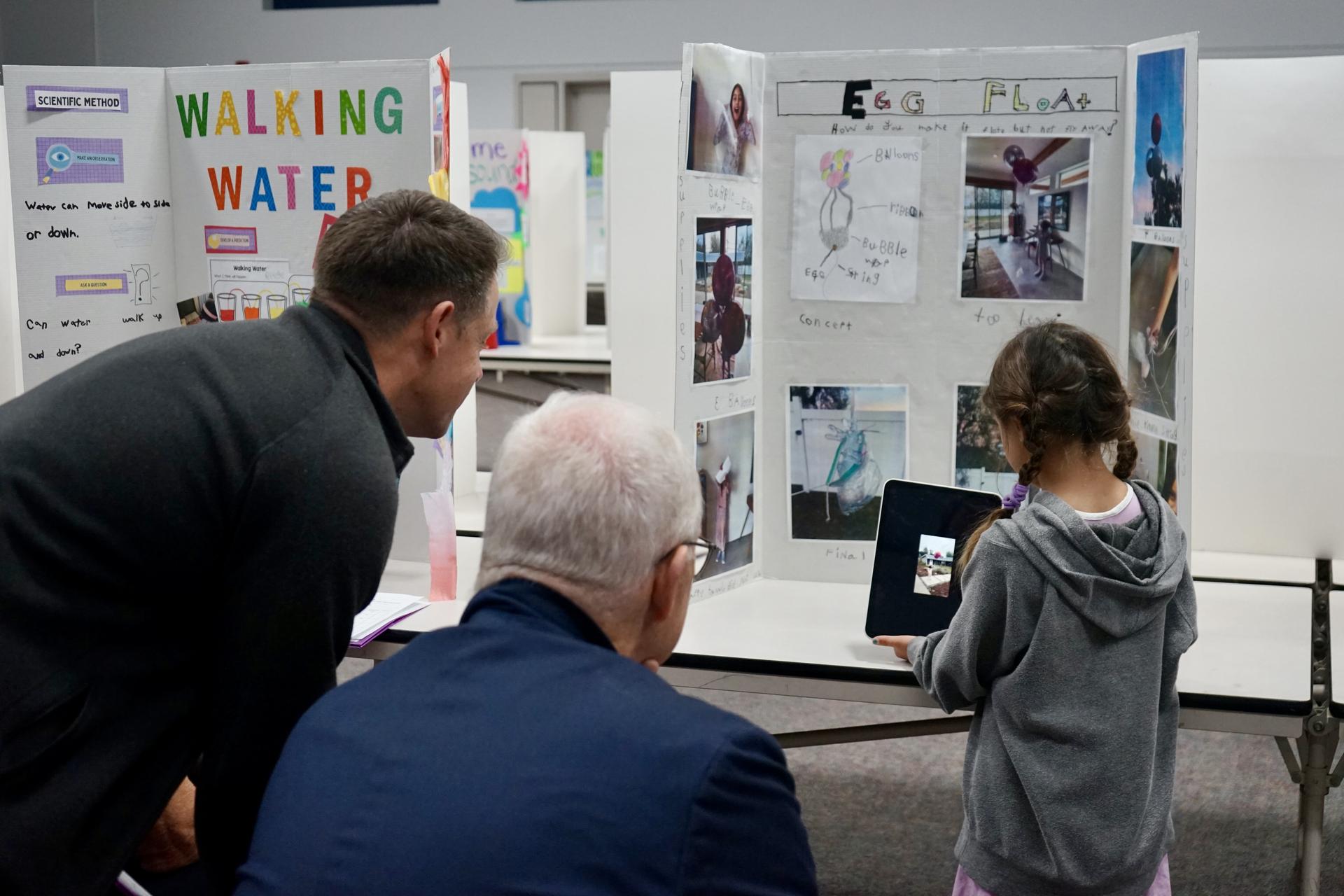 Two adult judges watch a video on an ipad held by a student as part of their science fair project.