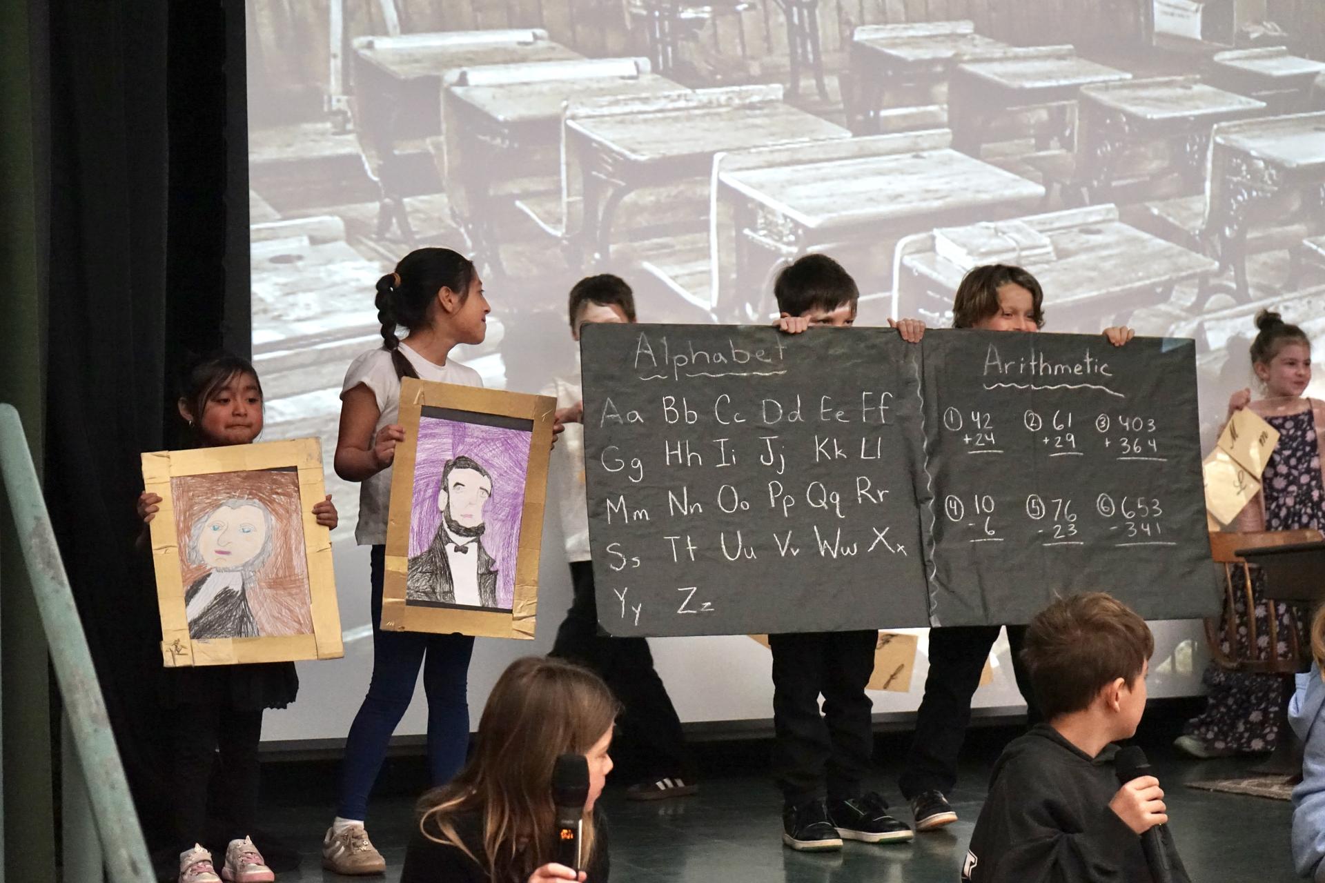 Five students stand on stage holding black posters meant to look like chalkboards.