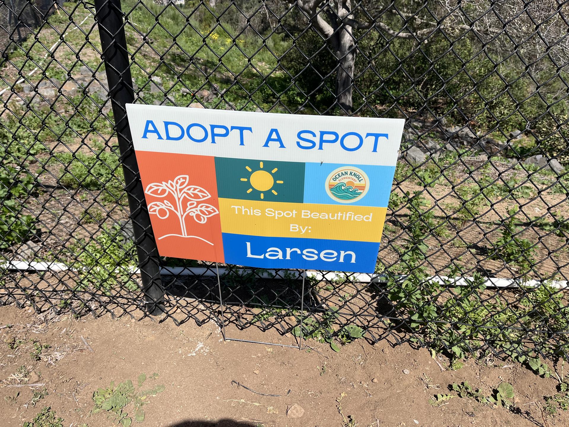 A sign attached to a fence that says Adopt A Spot, This spot beautified by: Larsen