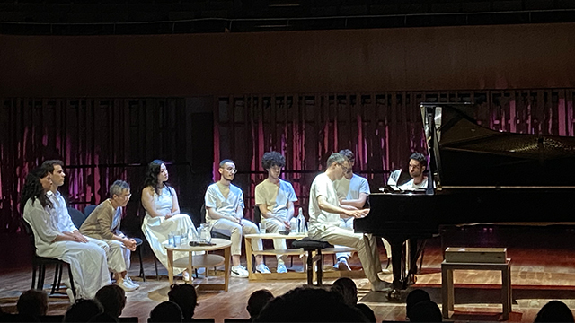 Rodolfo Faistauer sits at a piano on a stage with a small group of people seated in chairs behind