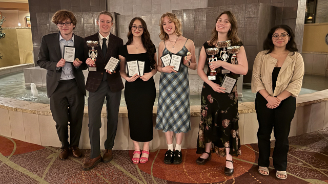 Six students, most holding plaques or trophies, stand in front of an indoor fountain