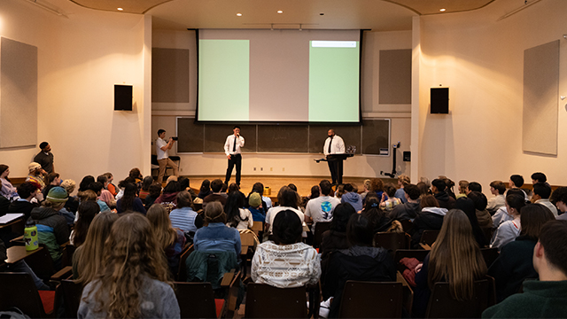 Two speakers on stage in Olin Auditorium; nearly every seat in the audience is filled