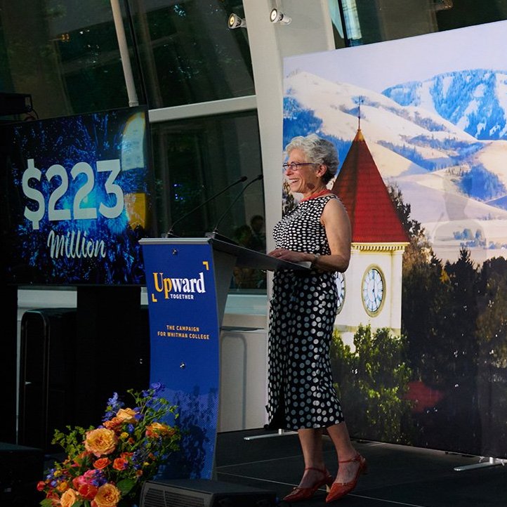 Nancy Serrurier stands at a podium behind an Upward Together sign with a screen in the background that reads ‘$223 Million.’