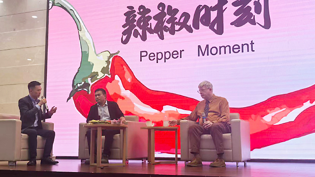 Brian Dott (right) on stage with two other presenters in front of a digital display of a large red pepper and the text “Pepper Moment”
