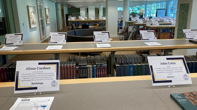 Library bookshelves with signs on top with the names of Whitman faculty members and information about their scholarly and creative work