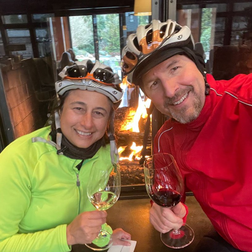 (selfie) Shilpa and Blake seated at a table in front of a fireplace holding wine glasses and wearing bicycle helmets