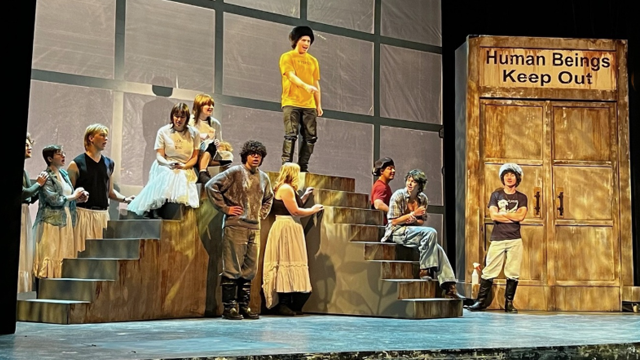A group of actors gathered by a set of stairs on a stage; on the right is a large closed door with a sign that reads “Human Beings Keep Out”