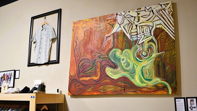A large art piece hangs on a wall next to a shirt on a hangar and some shoes on a shelf