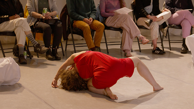 A dancer in red contorts their body on the floor with the legs of audience members visible in the background