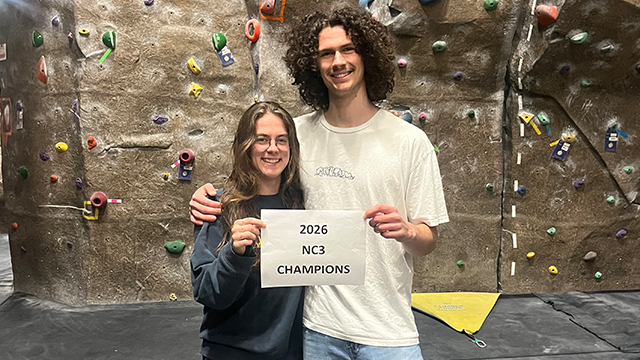 Lily Iseminger (left) and Orion Killian (right) stand in front of a climbing wall holding a white sheet of paper with the words “2026 NC3 Champions”