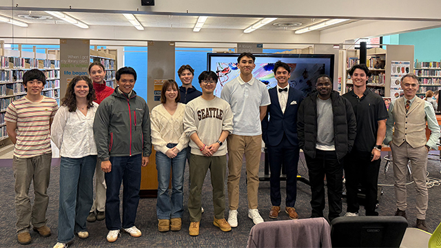 A group of students and Professor Andrés Aragoneses (far right) stand in front of rows of books at the library