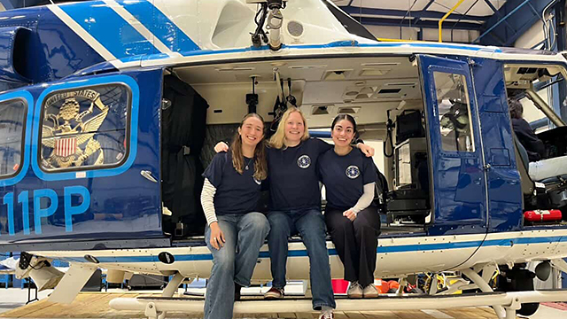 Three students wearing matching dark blue shirts and sitting on the edge of a helicopter in a hangar.
