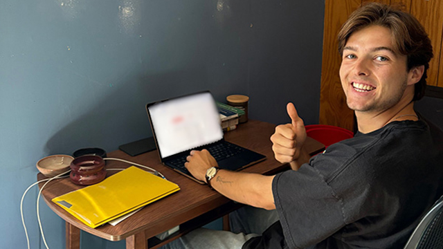 Gage Hembree sits at a small table with a laptop, smiling at the camera