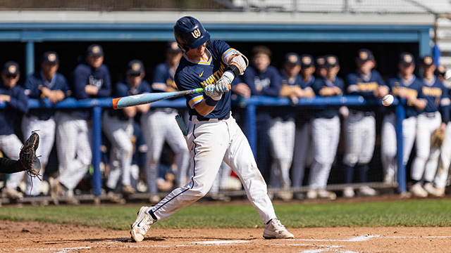 A Whitman baseball player swings at the ball with a line of teammates visible in the background