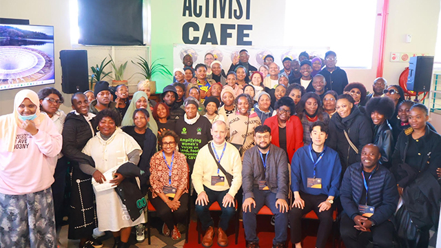Shampa Biswas (front row, fifth from right) and a large crowd of people pose for a photo in front of a sign that reads “Activist Cafe”