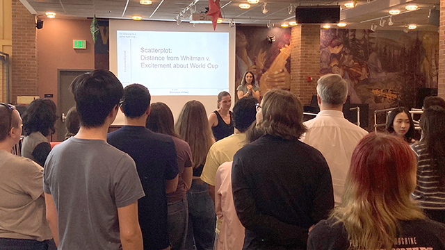 A group of people stand in the Reid Campus Center basement while Jessica Sutter stands in front of a screen that reads “Scatterplot: Distance from Whitman v. Excitement about World Cup”