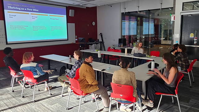 Students sit around a u-shaped group of tables in an office/classroom setting