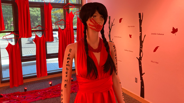 Red light illuminates a mannequin with a red handprint painted over the mouth; several red dresses hang in the background