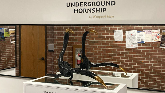 A black and gold horn shaped piece of art sits on a pedestal in front of a mirror and under text that reads: Underground Hornship by Wangechi Mutu