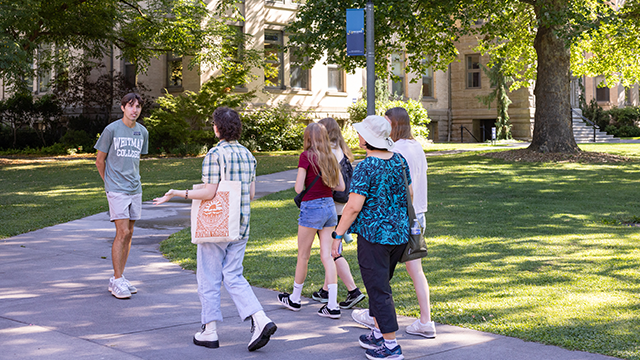 A student wearing a Whitman College shirt walks with a group of visitors by Memorial Hall