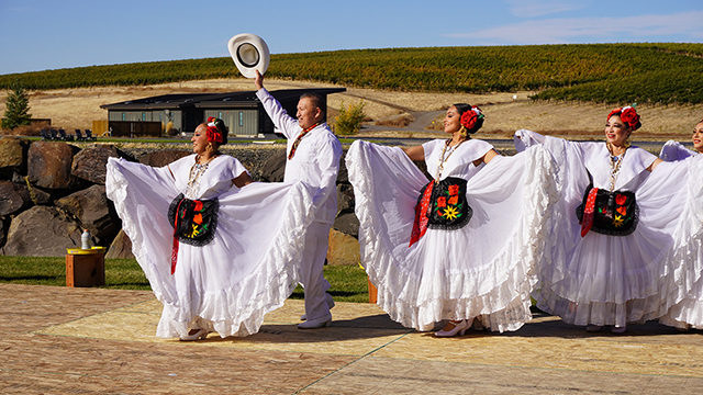 A group of dancers in white outfits perform outdoors with vineyards in the background