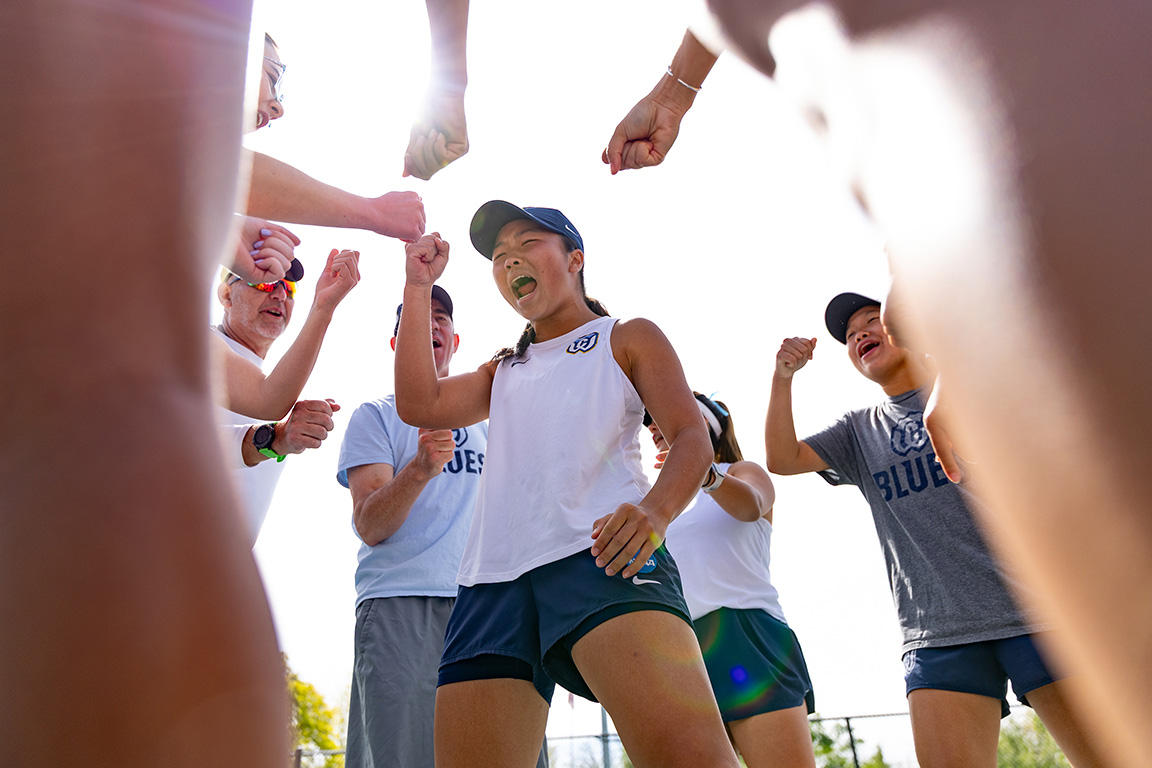 A Whitman tennis player stands in the center of a circle of coaches and athletes cheering.