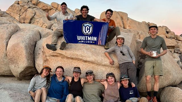 A group of people pose for a photo on a pile of large boulders; one person holds a blue “Whitman Blues” banner 