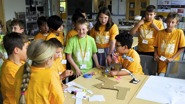 A group of elementary school children wearing “Kids College” t-shirts gather around a table with crafting materials