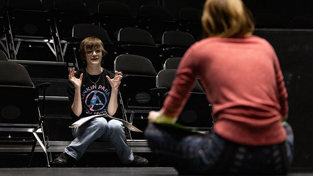 Two students with scripts in their laps talking in a theater rehearsal space