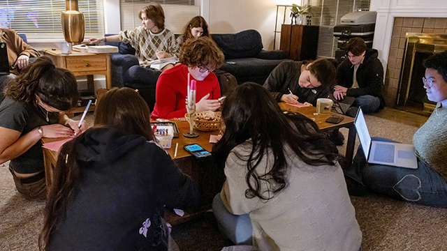 A group of students gather in a living room area, each working on a writing project