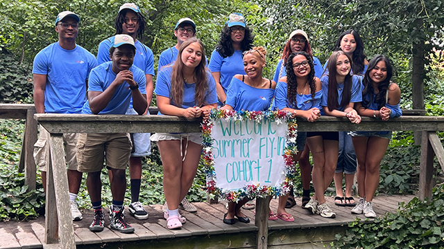 Twelve students in matching blue shirts stand on a wooden bridge with a sign that reads “Welcome Summer Fly-In Cohort 1”