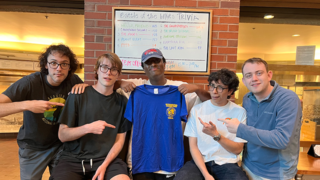 Five students stand in front of the Battle of the W(h)its Trivia results board