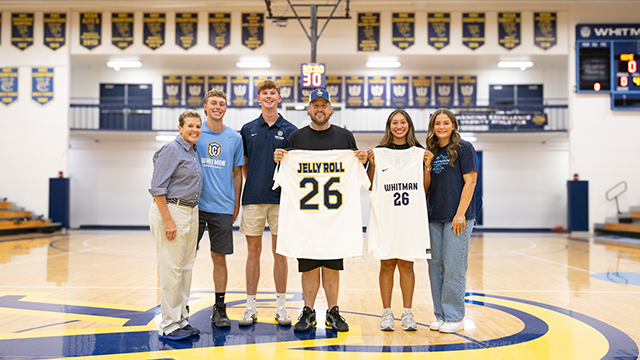A group of Whitman students and staff stand on a basketball court with Jelly Roll, holding up a custom Whitman jersey