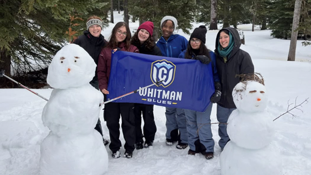 Six students in winter gear hold a “Whitman Blues” flag behind two snowmen.