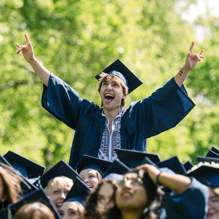 A graduate in a blue cap and gown celebrates above a crowd of classmates