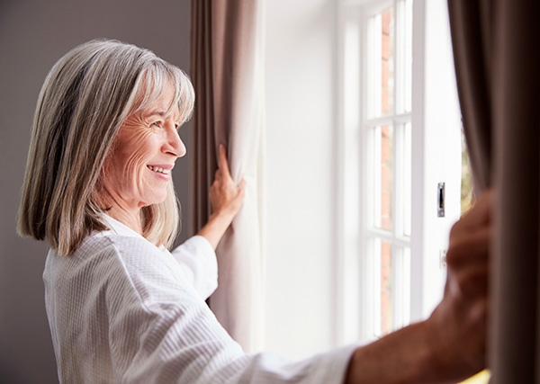 Smiling woman opening curtains and looking out window.