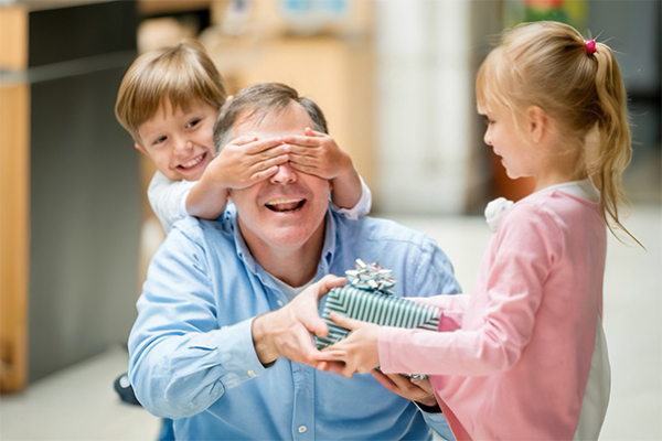 A young girl hands a present to an older man, while a young boy covers the man's eyes with his hands.