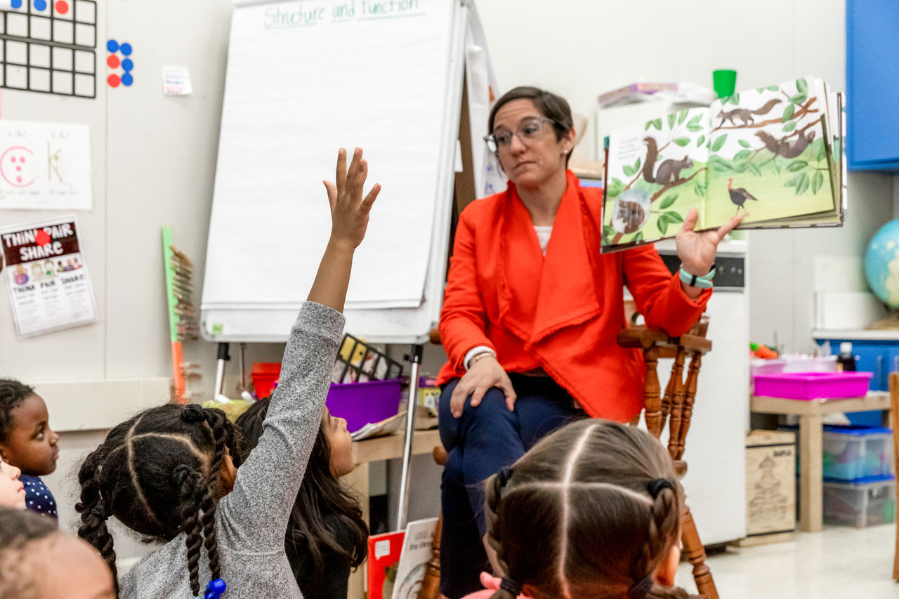 A first grade teacher reads to her students, one of whom has her hand raised