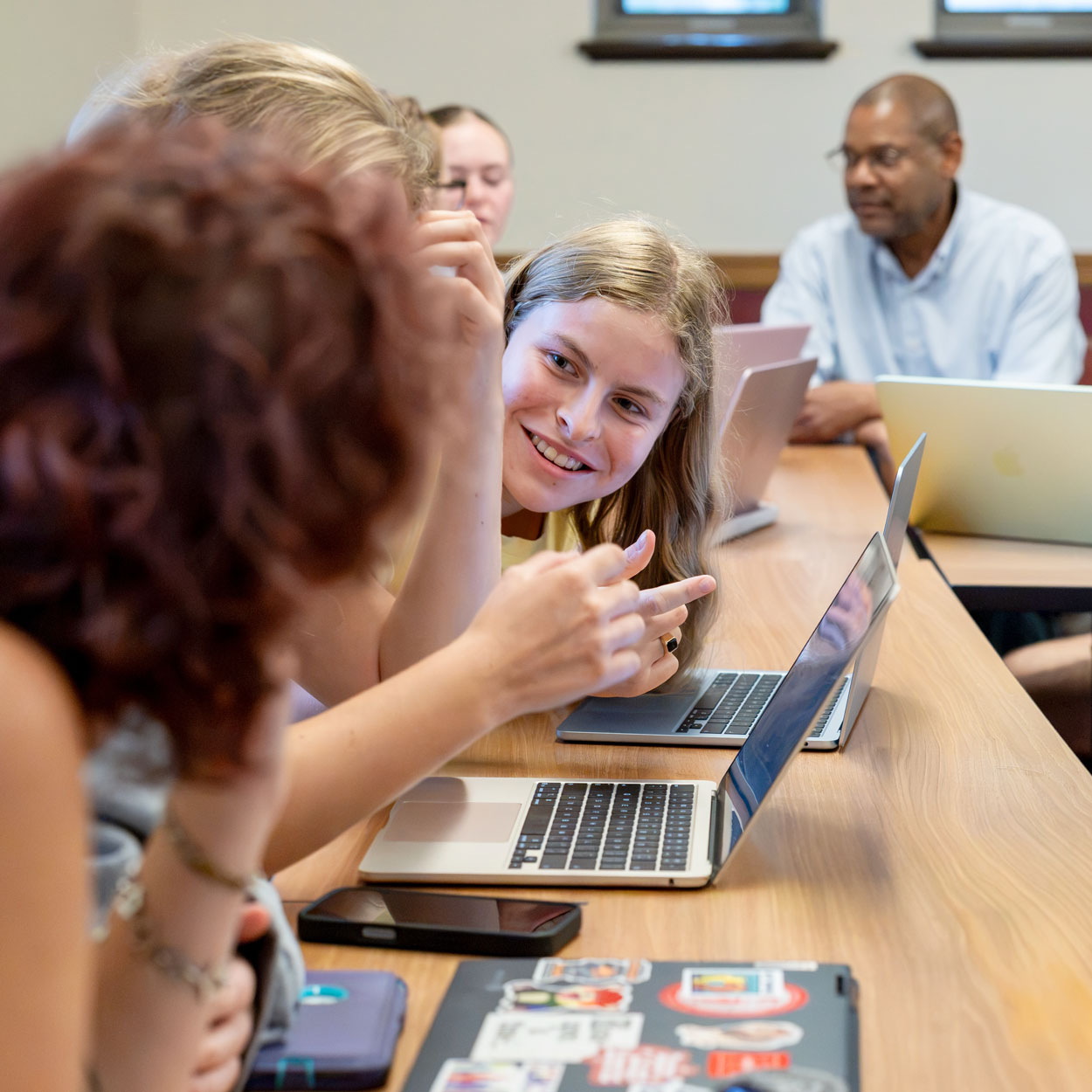 Three students sit at a table speaking in front of laptops, while the professor speaks to another student in the background