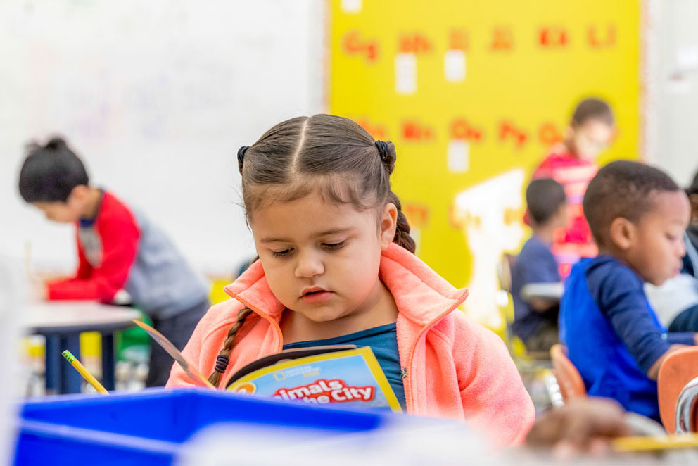A first grader reads in class while fellow students behind her complete schoolwork