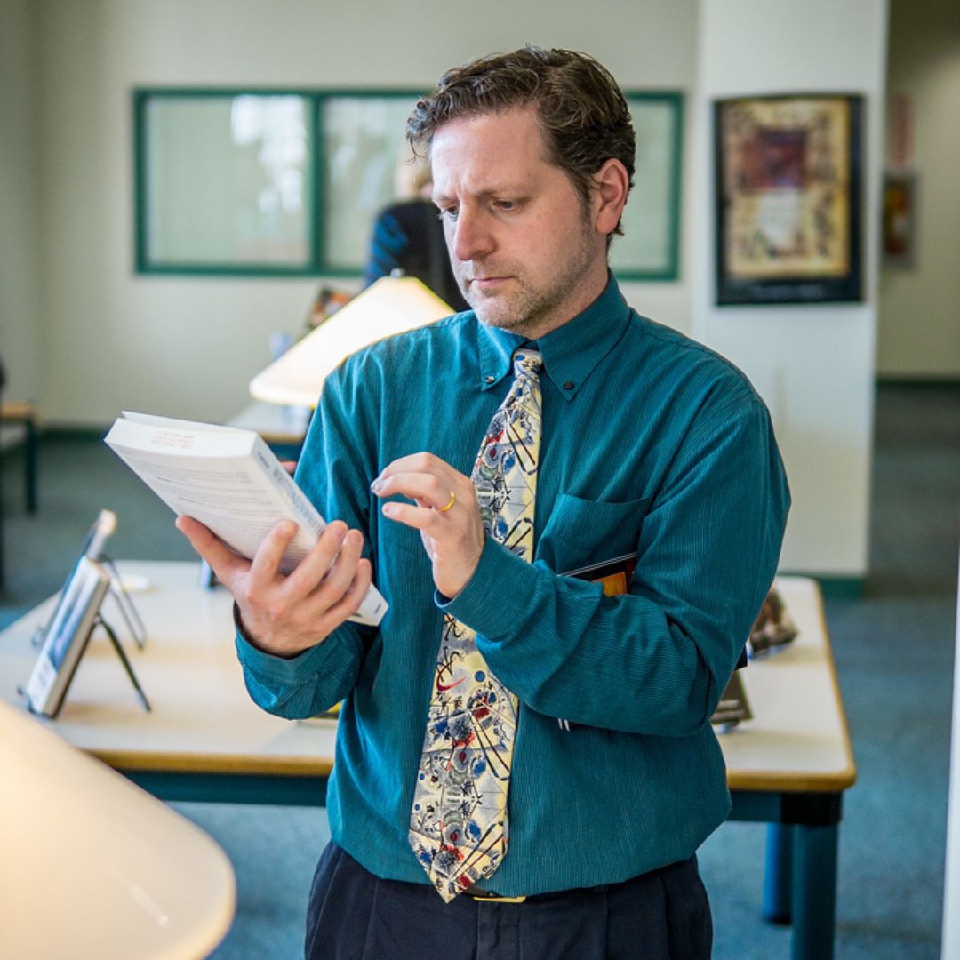Student holding a book between library shelves looking up at a higher shelf.