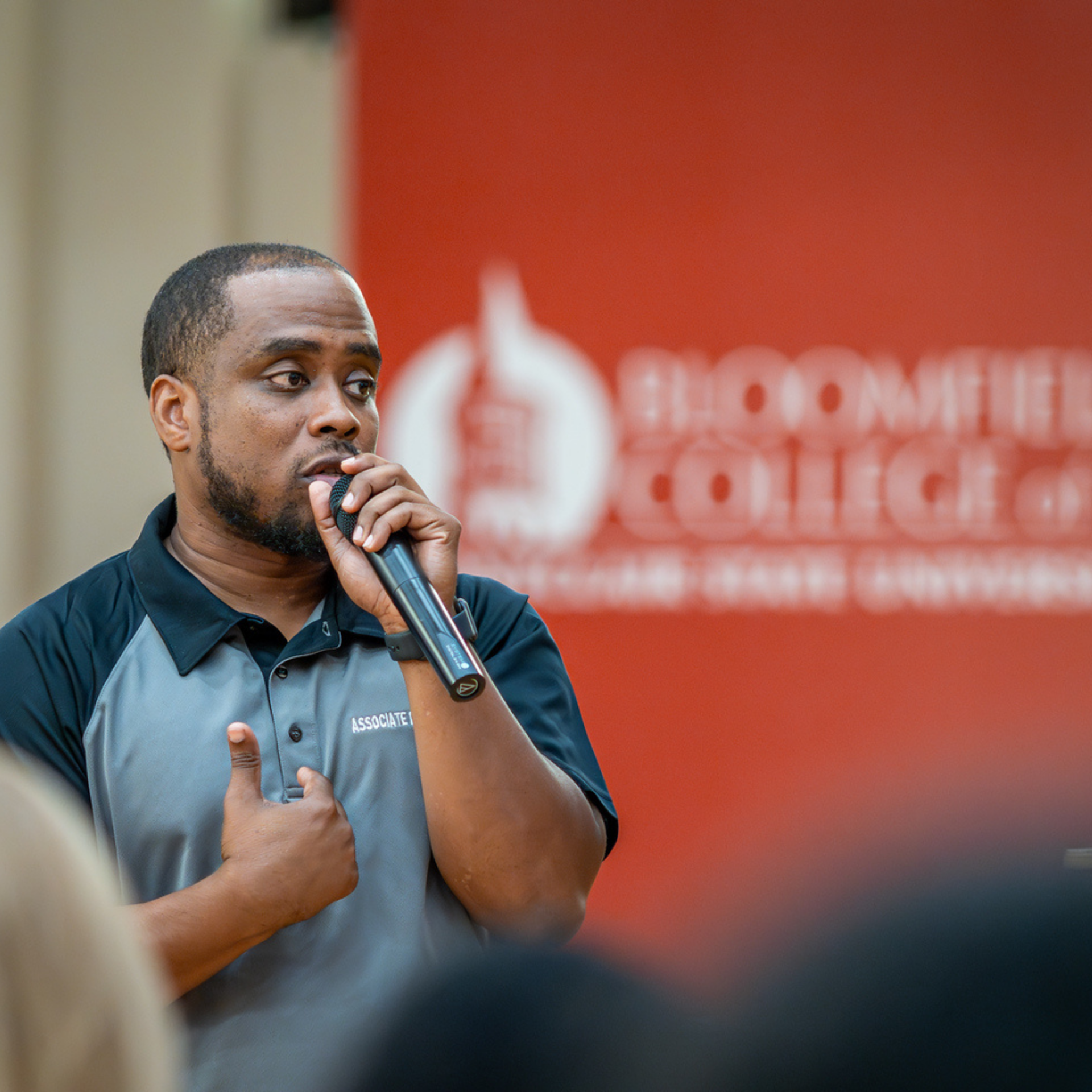 Staff member speaking into a microphone with a red Bloomfield College backdrop in the background.