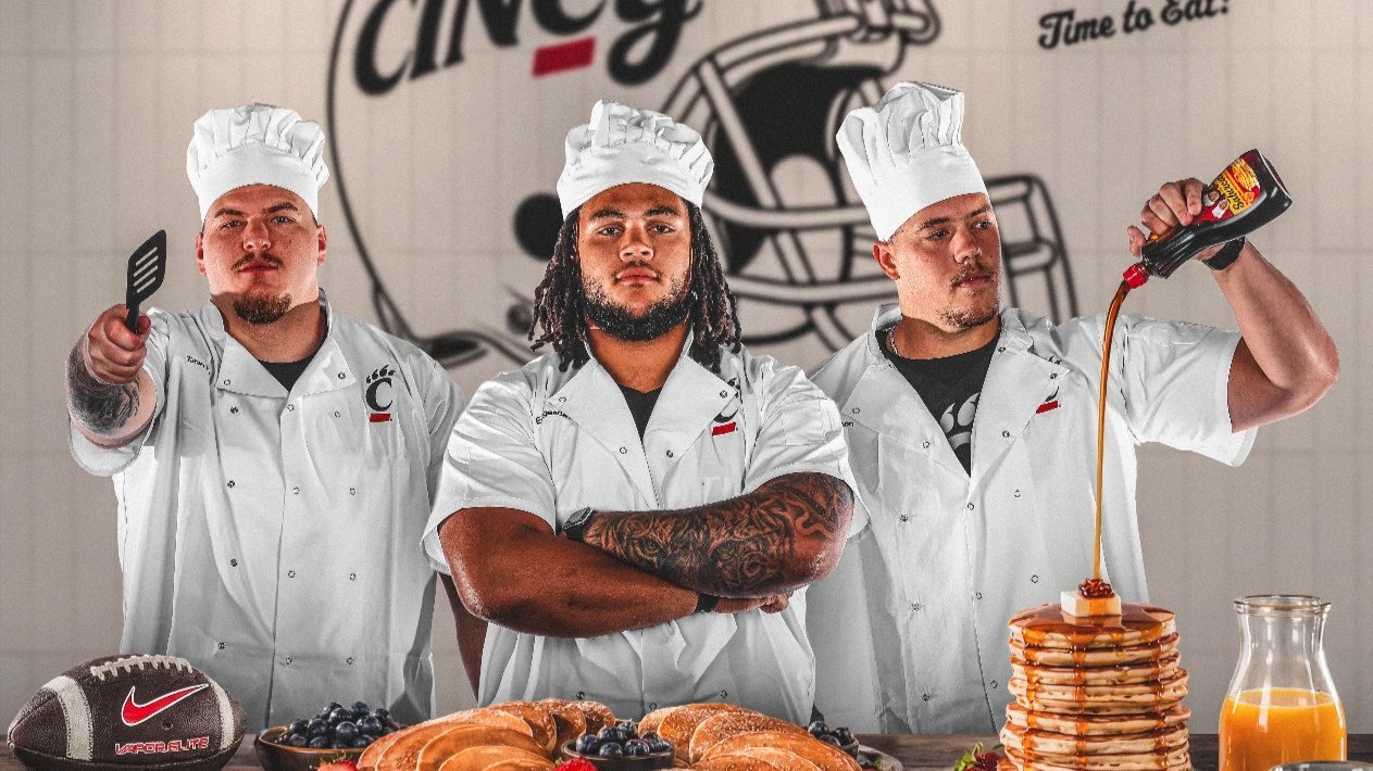 Three Cincinnati football players dressed as chefs pose behind a breakfast spread, with one pouring syrup over a stack of pancakes.